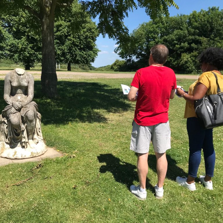 Psyche and survey A bright day in parkland. Headless seated statue to left of frame, mixed couple to right look at statue and hold paper.